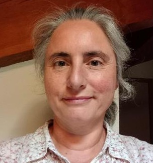 Portrait d’Isabelle Casenave, femme souriante aux cheveux gris, portant une chemise à motifs clairs, dans un intérieur chaleureux avec fond en bois, qui a participé à un atelier LEGO® SERIOUS PLAY® animé par les fondateurs de Trèfle Aventure